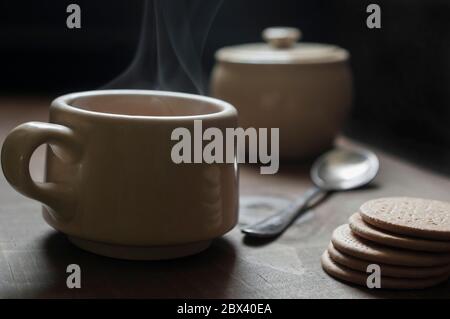 Una tazza di bevanda calda con un mucchio di biscotti sul lato e un cucchiaio sfocato e zuccheriera sullo sfondo, fotografia di cibo scuro Foto Stock
