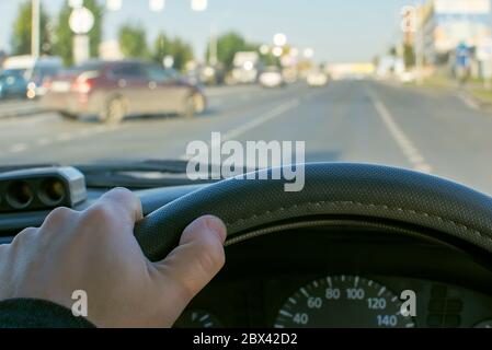 vista della mano del conducente sul volante di una macchina sullo sfondo di una strada urbana e di una macchina che lascia la strada secondaria sulla strada principale Foto Stock