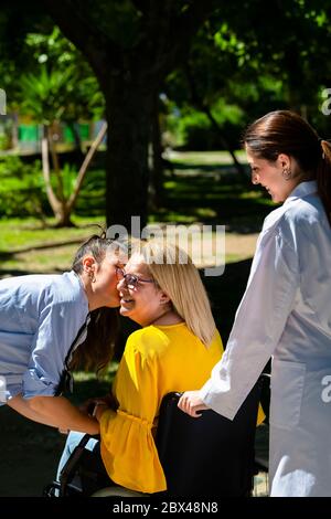 Una donna matura in sedia a rotelle sorride mentre viene baciata sulla guancia da un'altra donna su uno sfondo fuori fuoco. Concetto di salute e amicizia. Foto Stock