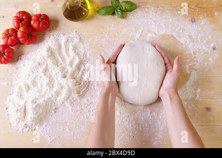 Vista dall'alto piatta delle mani dello chef donna che impastano la pasta per pizza sul piano di lavoro in legno e sulla farina, l'olio extra vergine, i pomodori e il basilico Foto Stock