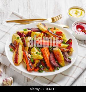 primo piano di barbabietole grigliate, spicchi di patate, pastinache, carote cosparse di cipolla verde tagliata su un piatto bianco con ketchup, senape, terreni Foto Stock