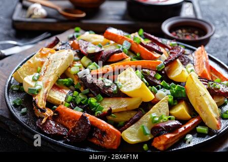 primo piano di barbabietole grigliate, spicchi di patate, pastinache, carote cosparse di cipolla verde tagliata su un asse di legno con ketchup, senape Foto Stock