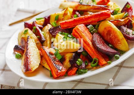 primo piano di barbabietole grigliate, spicchi di patate, pastinache, carote cosparse di cipolla verde tagliata su un piatto bianco con ketchup, senape, oriz Foto Stock