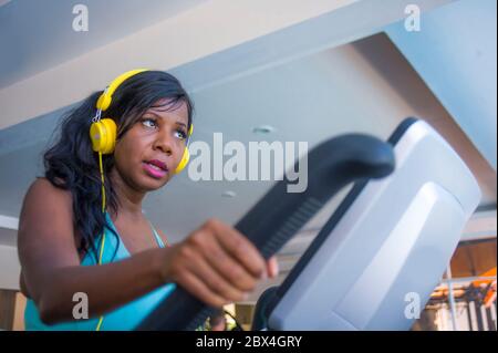 All'interno ritratto palestra di giovane attraente e concentrata donna afroamericana nera con cuffie allenamento ellittico macchina allenamento al fitness clu Foto Stock