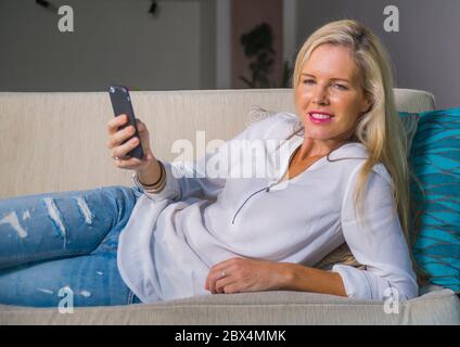 bella e felice bionda donna all'inizio degli anni '40 si rilassò nel salotto di casa utilizzando l'app social media su internet sul telefono cellulare sorridendo sdraiato comodamente su sof Foto Stock