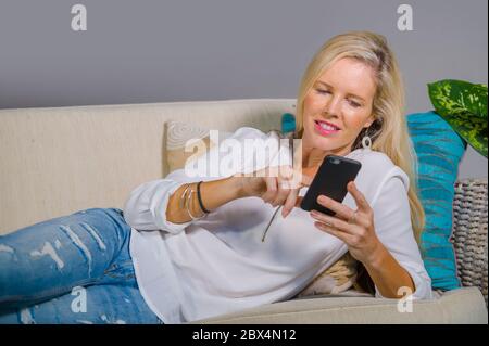 bella e felice bionda donna all'inizio degli anni '40 si rilassò nel salotto di casa utilizzando l'app social media su internet sul telefono cellulare sorridendo sdraiato comodamente su sof Foto Stock