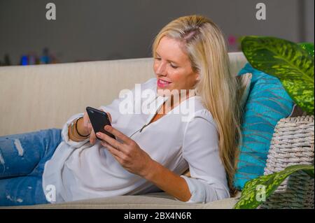 bella e felice bionda donna all'inizio degli anni '40 si rilassò nel salotto di casa utilizzando l'app social media su internet sul telefono cellulare sorridendo sdraiato comodamente su sof Foto Stock