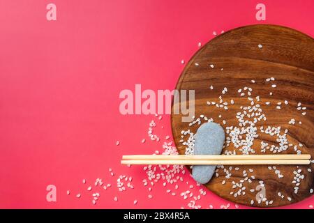 Disposizione piatta. I bastoncini di legno chiaro giacciono su pietre ovali e su una superficie di legno su sfondo rosa. Il riso è sparso casualmente sulla superficie. Il Foto Stock