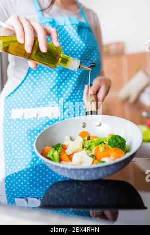 Donna che versa olio d'oliva sulle verdure in padella a casa. Concetto di pasto sano. Foto Stock