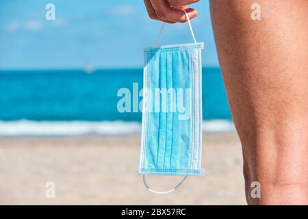 primo piano di un giovane caucasico, visto da dietro, in piedi sulla spiaggia, tenendo in mano una maschera chirurgica blu, come lo è prendere un respiratore di vestibilità Foto Stock