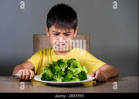 7 o 8 anni sconvolto e disgustato bambino asiatico seduto sul tavolo di fronte al piatto di broccoli sembra infelice rifiutare il cibo fresco in bambino gele odio Foto Stock