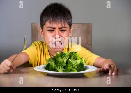 7 o 8 anni sconvolto e disgustato bambino asiatico seduto sul tavolo di fronte al piatto di broccoli sembra infelice rifiutare il cibo fresco in bambino gele odio Foto Stock