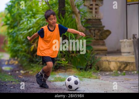 8 o 9 anni felice ed eccitato capretto che gioca il calcio all'aperto in giardino che indossa il giubbotto di addestramento che corre e calcia la sfera di calcio, il capretto che ha divertimento Foto Stock