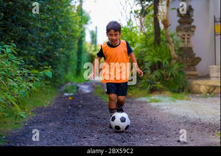 8 o 9 anni felice ed eccitato capretto che gioca il calcio all'aperto in giardino che indossa il giubbotto di addestramento che corre e calcia la sfera di calcio, il capretto che ha divertimento Foto Stock