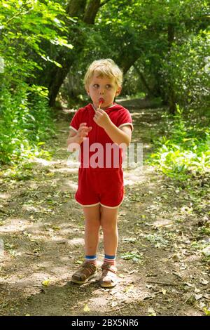 Ragazzo di tre anni in un vestito rosso da gioco che mangia un lollypop, Hampshire, Inghilterra, Regno Unito. Foto Stock