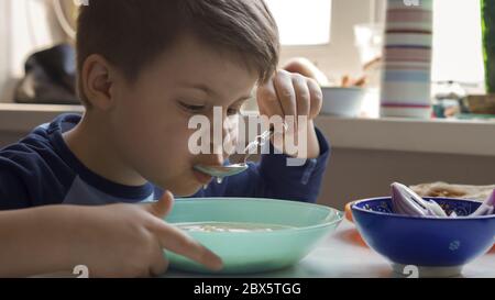 Un ragazzo allegro mangia la zuppa con un cucchiaio mentre si siede al tavolo da cucina. Pasti affamati di preschooler a casa Foto Stock