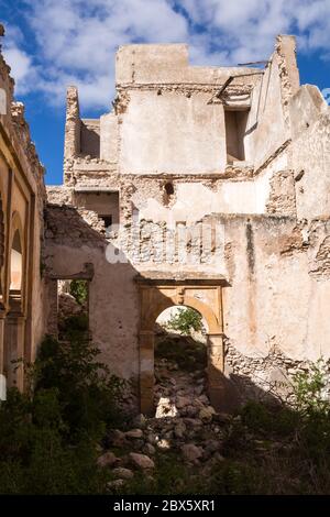 Vecchio edificio abbandonato in rovina. Cancello con arco senza porta. Rovinata facciata delle pareti. Cespugli selvaggi. Cielo blu con nuvole bianche. Dar Caid H Foto Stock