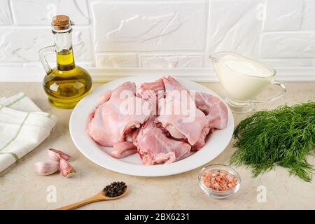 Pezzi di carne di coniglio cruda, panna acida, aglio, pepe, sale, olio d'oliva e mazzo di aneto sul tavolo da cucina. Ingredienti per cucinare stufato di coniglio. Foto Stock