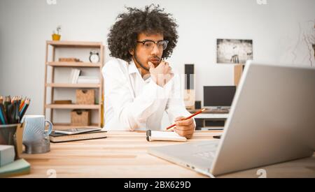 Il giovane pensivo è impegnato a casa davanti al computer. Studente arabo o uomo d'affari che prende appunti in un notebook. Concetto di educazione domestica. Immagine a toni Foto Stock