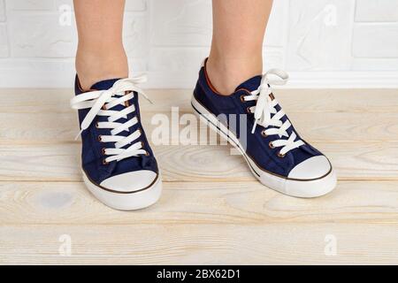 I piedi della donna si stacchettano in scarpe da ginnastica classiche blue con lacci bianchi e si levano in piedi sui piedi su un pavimento di legno bianco. Indossa le scarpe sportive da vicino. Foto Stock