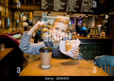 Divertente adorabile ragazzo seduto al tavolo in caffetteria, mangiando croissant fresco e mostrando la sua forza tenendo il braccio piegato al gomito Foto Stock