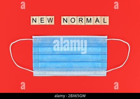 Piatto con maschera protettiva monouso blu e lettere in legno che formano "nuovo normale" su sfondo rosso Foto Stock