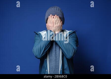 Uomo caucasico in un cappello a maglia e giacca impermeabile coperto di faccia con le mani, nascondendosi da problemi, su uno sfondo blu. Foto Stock