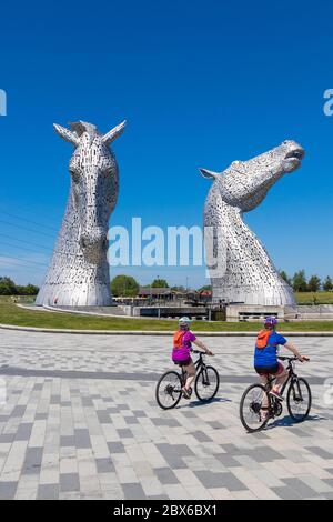 Due ciclisti in bicicletta davanti alle massicce sculture di cavalli chiamate Kelpies, vicino Falkirk, Scozia Foto Stock