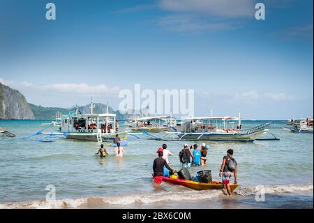 Turisti e le mani locali della barca filippina si sfilano o portano rifornimenti alle numerose barche ormeggiate nel surf in attesa di visitare le isole e le spiagge. Foto Stock