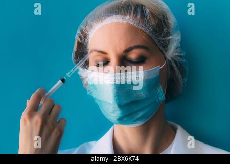 Primo piano ritratto di giovane dottoressa in cappuccio medico e maschera, abito bianco e siringa sulla sua mano. Medico con occhi chiusi stanco dopo l'iniezione Foto Stock