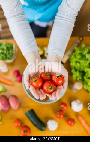 Vista sopra di donna che indossa grembiule che tiene piccoli pomodori con verdure colorate su tavolo da cucina. Sana preparazione dei pasti fatti in casa. Foto Stock