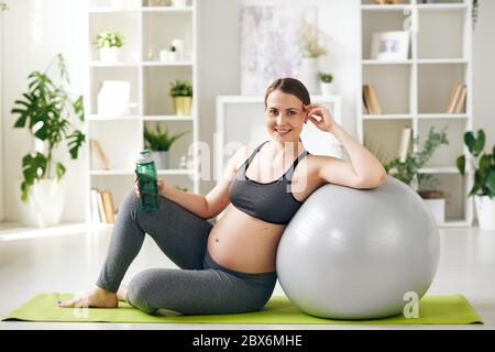 Giovane donna incinta sportiva di successo con una bottiglia d'acqua appoggiata al pallone da fitball mentre si siede sul tappetino in un ambiente domestico Foto Stock