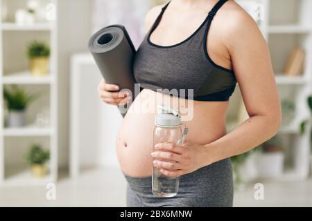 Giovane donna incinta attiva con tappetino arrotolato e bottiglia di acqua pura ferma in piedi davanti alla macchina fotografica mentre si va fare esercizi fisici Foto Stock