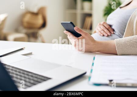 Mano di giovane donna d'affari incinta che usa lo smartphone mentre si siede alla scrivania davanti al computer portatile in ufficio e in ufficio Foto Stock