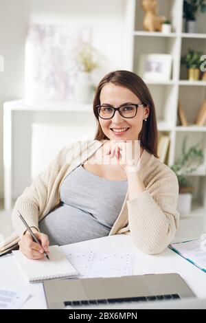 Giovane manager incinta di successo che analizza grafico finanziario e prende appunti in blocco note mentre lavora davanti alla fotocamera in ufficio Foto Stock