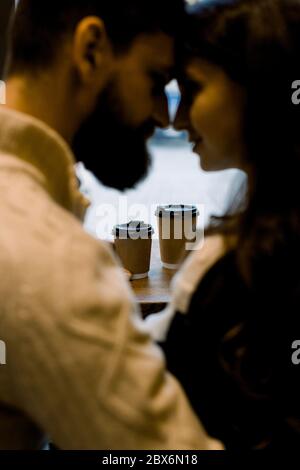 Primo piano ritratto di giovane coppia romantica che beve caffè in tazze di carta, seduto in un accogliente caffè, toccando le fronte e godendo momenti insieme Foto Stock