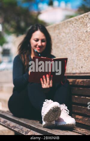 Giovane donna sfocata si siede sulla panchina nel parco con notebook e penna. Wish Marathon. Scrivere desideri e sogni in un notebook. Vita con un'ardesia pulita. Nuovo Foto Stock