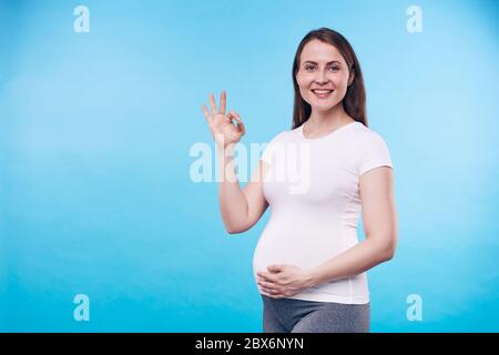 Felice giovane donna incinta mostrando il segno giusto mentre la tocca la pancia e guardandola in isolamento contro sfondo blu Foto Stock