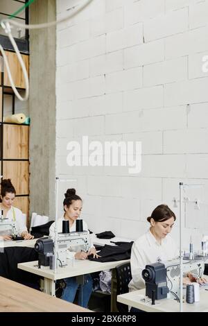 Fila di giovani stilisti seduti da scrivanie e utilizzando macchine da cucire elettriche mentre si lavora su nuova collezione stagionale Foto Stock