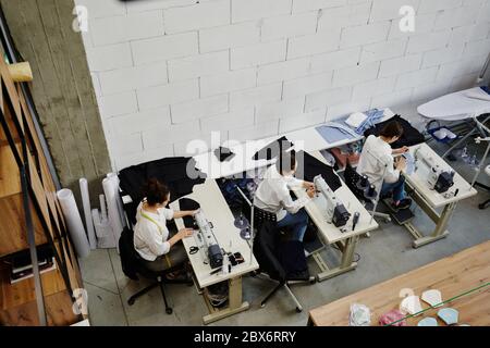 Panoramica di tre sarti seduti su una scrivania con macchine elettriche per cucire durante il lavoro su una nuova collezione di moda in officina Foto Stock