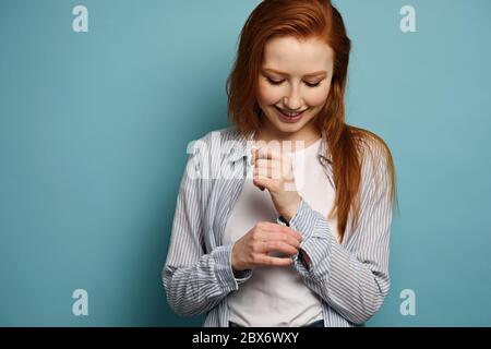 Bella ragazza dai capelli rossi in una camicia a righe si erge su uno sfondo blu sorridente e che abbellita un bottone su un bracciale. Foto Stock