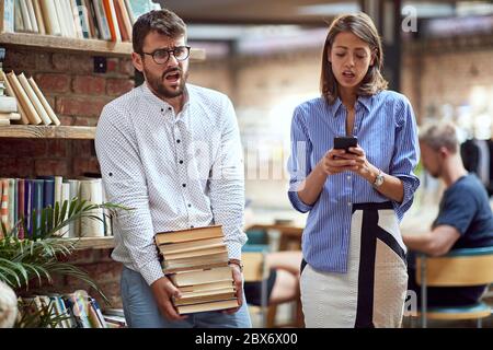 vecchio-fashioned maschio che porta un mucchio pesante di libri alla biblioteca, lottando. femmina moderna che guarda i e-libri sul suo telefono delle cellule. libri contro il concetto di e-libri Foto Stock