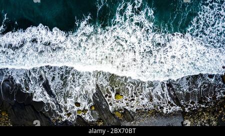 Vista aerea sulle onde che si infrangono con pietre colorate sulla spiaggia di sabbia nera. Sfondo astratto. Acque verdi scure. Foto Stock