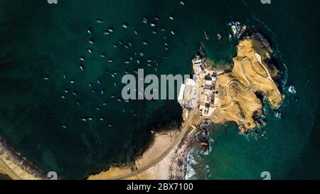 Vista aerea di molte barche da pesca vicino alla penisola sulla costa rocciosa del Perù. Piccole barche da pesca dall'alto. Acque verde scuro, isola rocciosa Foto Stock