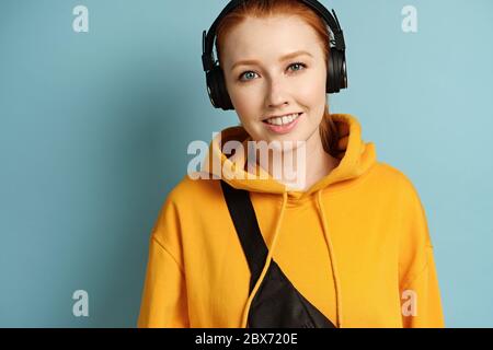 Ragazza dai capelli rossi in una felpa gialla e cuffie si erge su uno sfondo blu e sorride Foto Stock