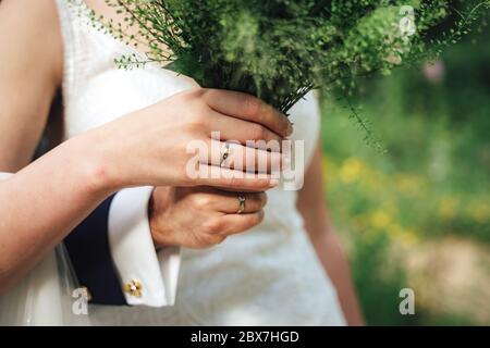 Vista in primo piano delle mani degli sposi nuovi con colorato bouquet di nozze. Sposa e sposo che indossano anelli da sposa. Sfondo esterno. Concetto di giorno di nozze. Foto Stock