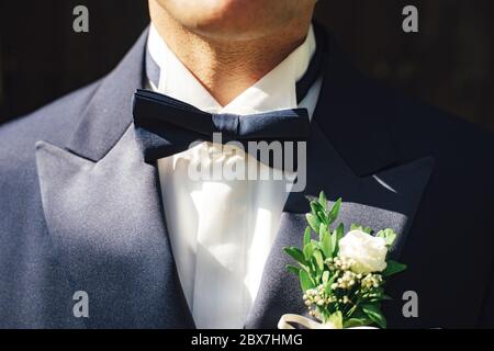 Primo piano di uomo in camicia bianca e vestito blu con cravatta di arco blu e fiore matrimonio boutonniere. Sfondo scuro. Foto Stock