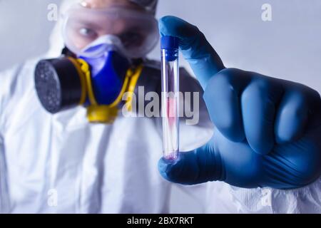 Laboratorio completamente attrezzato in respiratore, occhiali, abbigliamento e guanti di gomma blu che tengono e guardano la provetta con virus su sfondo colorato. Foto Stock