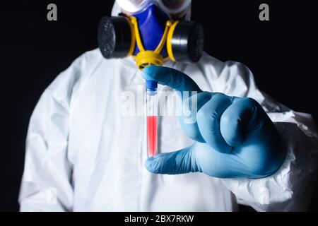 Laboratorio completamente attrezzato in respiratore, occhiali, abbigliamento e guanti di gomma blu che tengono una provetta con virus su sfondo nero. Foto Stock