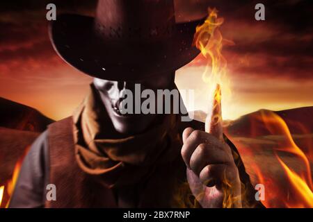 Foto di un cowboy demoniaco del cranio in cappello che getta incantesimo di fuoco con il suo braccio e dito su uno sfondo desertico tramonto. Foto Stock